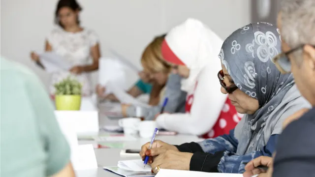 Vrouw aan tafel met papierwerk samen met andere vluchtelingen