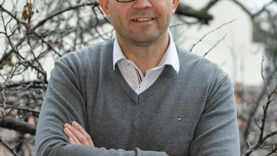 Een man met een zwarte bril, grijze trui en witte kraag heeft zijn armen over elkaar en poseert met een glimlach. De achtergrond bestaat uit de lege takken van een boom.