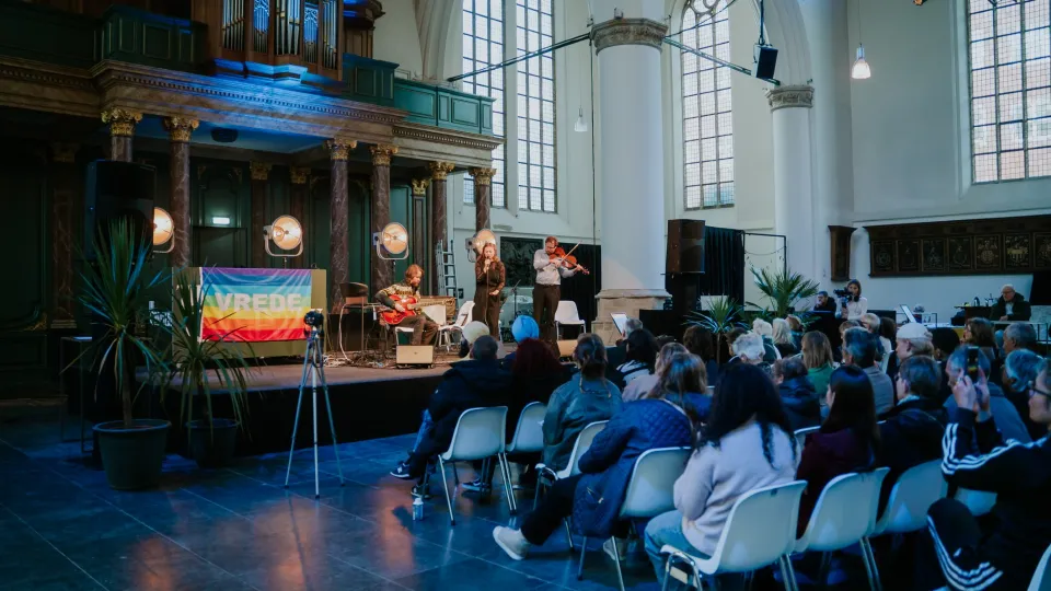 muzikanten op het podium in de Grote kerk in Den Haag en er is publiek aanwezig. Regenboogvlag met het woord 'VREDE'.