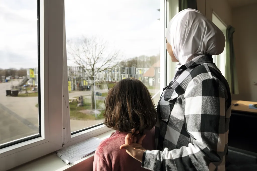 Een moeder en kind kijken uit het raam in een asielzoekerscentrum