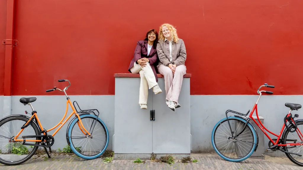 Rachel en Esmee zitten tegen een rode muur aan. 